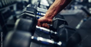 A muscular hand reaches for a heavy dumbbell from a full weight rack inside a gym, with more dumbbells in the background.