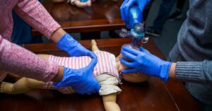 Two providers in blue gloves perform bag-mask ventilation on a pediatric mannequin during hands-on PALS certification training.