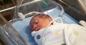 A newborn infant rests in a hospital bassinet under an oxygen hood, swaddled in a white blanket with colorful dots.