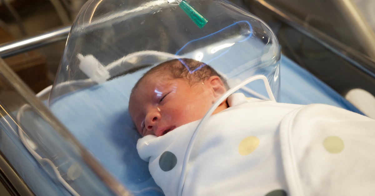 A newborn infant rests in a hospital bassinet under an oxygen hood, swaddled in a white blanket with colorful dots.