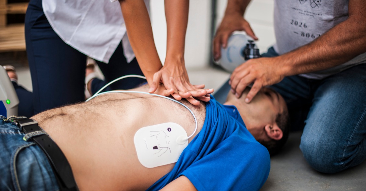 A man on the ground with his shirt pulled up, with defibrillator patches on his chest, while a woman performs CPR.
