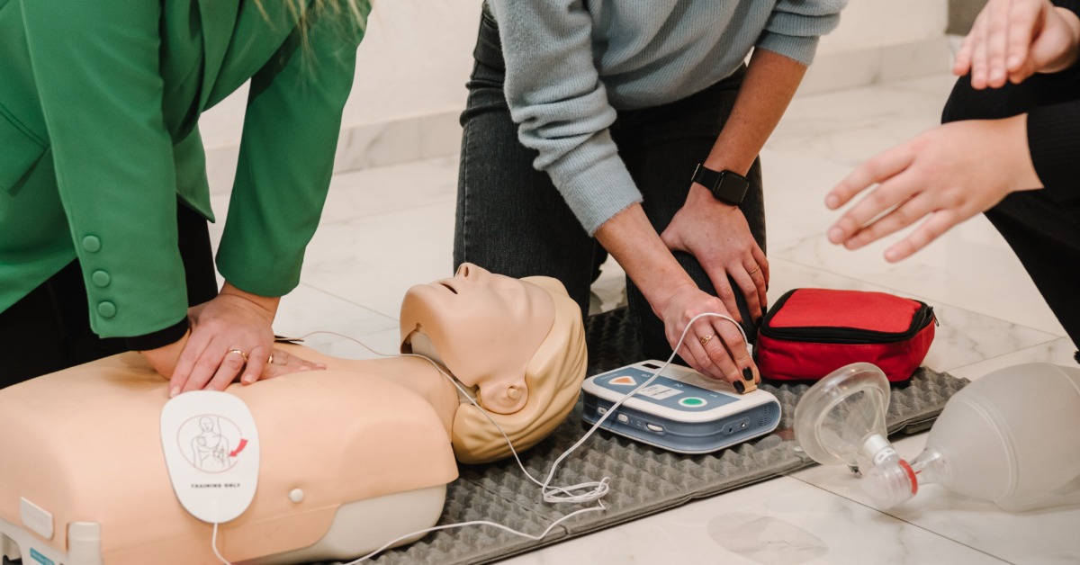 A close-up of a CPR training dummy on the floor as three women practice using an external defibrillator device.