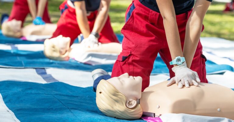 Trainees in red uniforms practice CPR chest compressions on mannequins during an outdoor class.